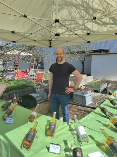Mann steht an einem Marktstand mit Flaschen von Getränken und grünem Tischdecken.